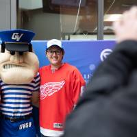 Two alumni, wearing Red Wings Jerseys pose with Louie the Laker at the Detroit Red Wings GVSU Night